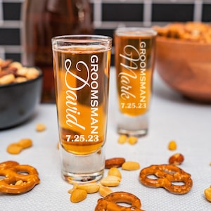 May include: Two clear glass shot glasses with the words "Groomman" and the date "7.25.23" etched on them. The glasses are filled with a light brown liquid and are sitting on a white surface with pretzels and peanuts.