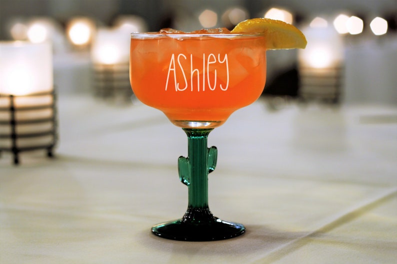 May include: A margarita glass with a cactus stem and a slice of lemon. The glass is filled with a pink drink and has the name "Ashley" written on it.
