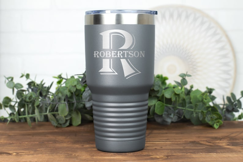 May include: A gray insulated tumbler with a silver rim and lid. The tumbler features the name "Robertson" and a large letter "R" etched in white. The tumbler has horizontal ridges and is set on a wooden surface.