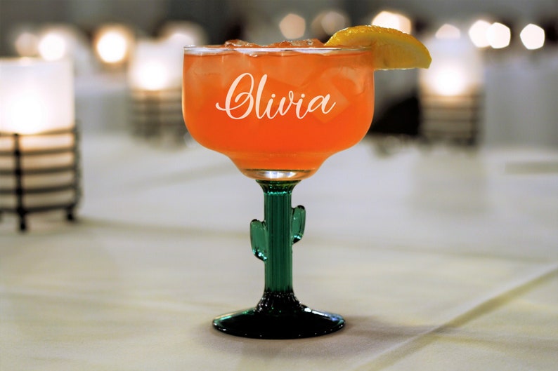 May include: A clear margarita glass with a green cactus stem and base. The glass is filled with a pink drink and a slice of lemon. The glass is personalized with the name "Olivia" in white lettering.
