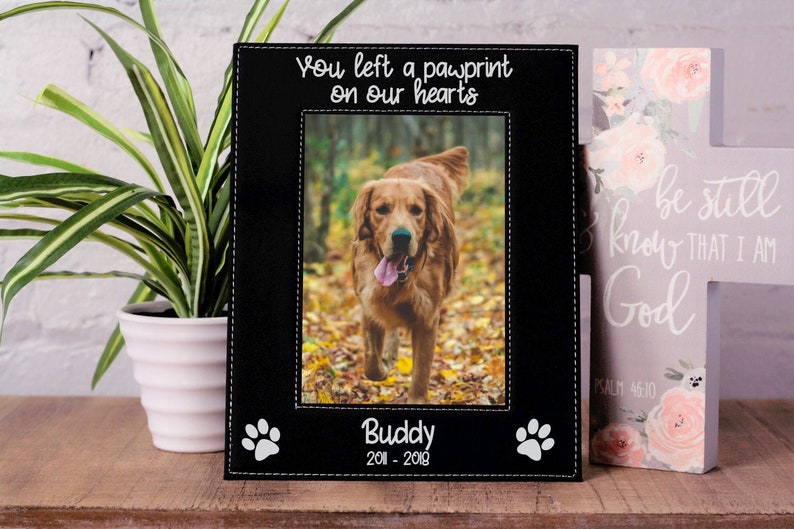 May include: Black leather photo frame with a stitched border and white paw prints. The frame has a photo of a golden retriever dog and the text "You left a pawprint on our hearts" and "Buddy 2011 - 2018".