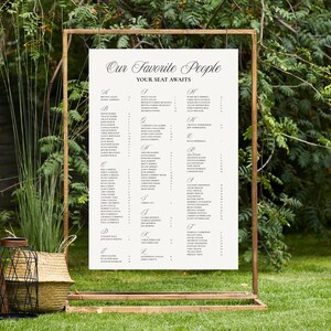 May include: A large seating chart with the text "Our Favorite People, Your Seat Awaits" is displayed on a wooden frame. The chart is cream-colored with black text, listing names alphabetically. The scene is outdoors, with greenery in the background.