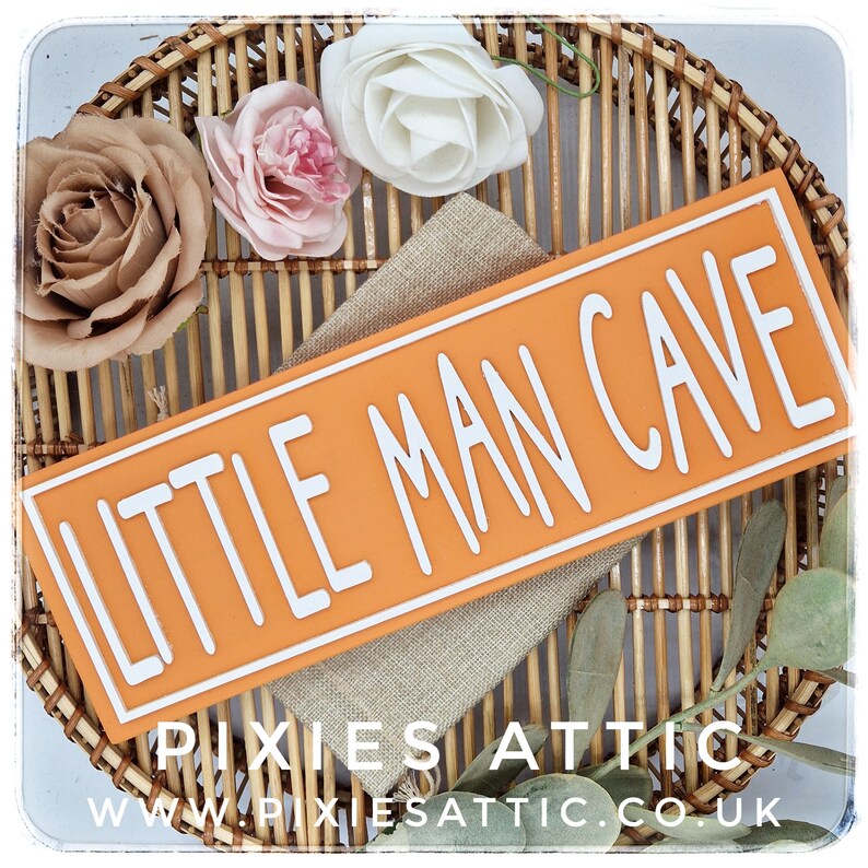 May include: An orange rectangular sign with the words "LITTLE MAN CAVE" in white lettering. The sign is set on a burlap cloth and surrounded by decorative flowers and greenery. The sign is displayed on a woven wicker tray.