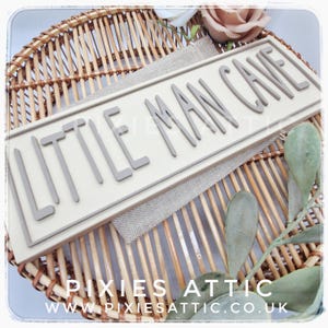 May include: A rectangular sign with the words "LITTLE MAN CAVE" in gray, raised letters. The sign is cream-colored and sits on a woven wicker basket with a burlap-like fabric. The sign is surrounded by decorative elements.