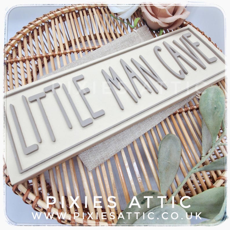 May include: A rectangular sign with the words "LITTLE MAN CAVE" in gray, raised letters. The sign is cream-colored and sits on a woven wicker basket with a burlap-like fabric. The sign is surrounded by decorative elements.