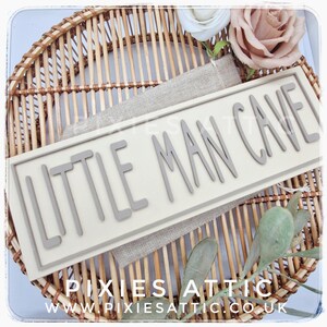 May include: A rectangular sign with the words "LITTLE MAN CAVE" in gray, raised lettering. The sign is set against a neutral background and rests on a woven wicker tray. Decorative flowers and greenery are also present.