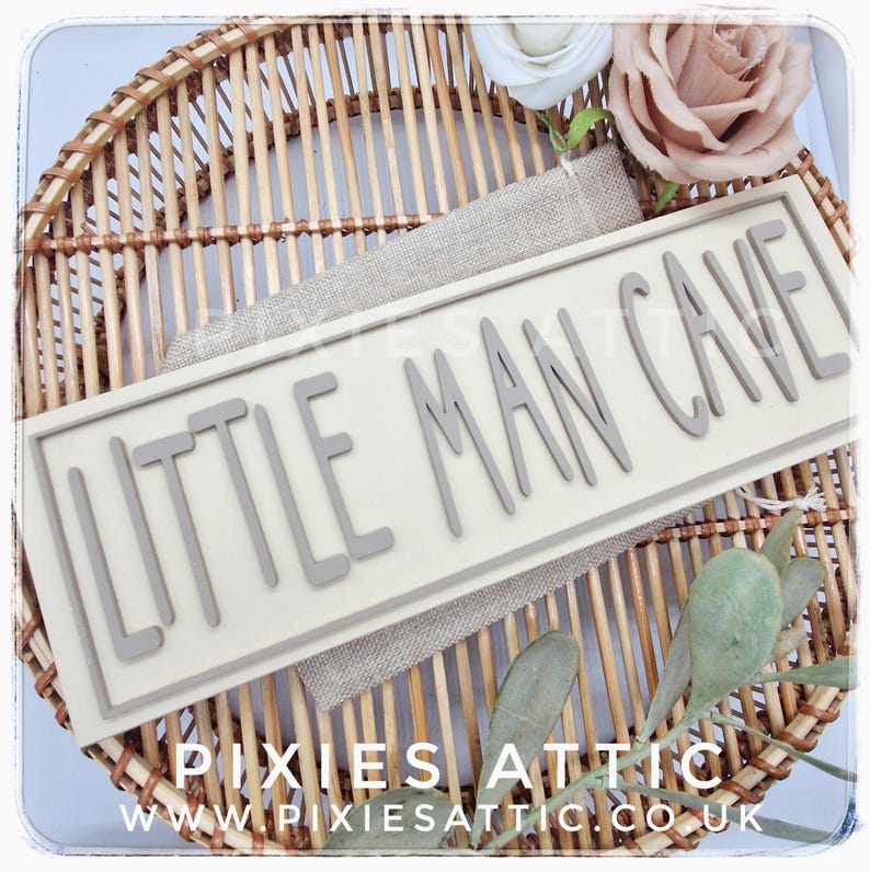 May include: A rectangular sign with the words "LITTLE MAN CAVE" in gray, raised lettering. The sign is set against a neutral background and rests on a woven wicker tray. Decorative flowers and greenery are also present.