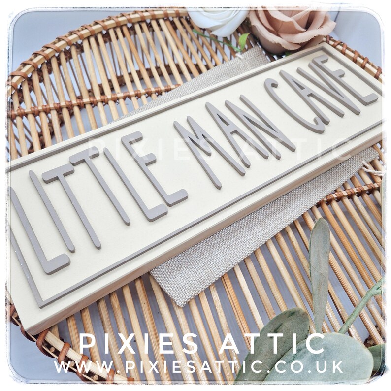 May include: A rectangular wooden sign with the words "LITTLE MAN CAVE" in gray, raised letters. The sign is set against a light beige background and framed in a light gray border. The sign is placed on a woven wicker tray.