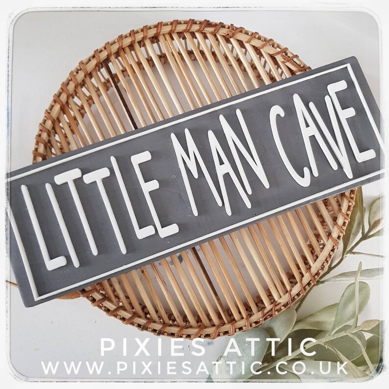 May include: A rectangular gray sign with the words "LITTLE MAN CAVE" in white, set against a woven basket background. The sign has a white border. The bottom of the image includes the text "PIXIES ATTIC" and the website address.