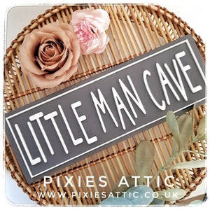 May include: A gray rectangular sign with the words "LITTLE MAN CAVE" in white, set against a white border. The sign is placed on a woven wicker tray, adorned with faux flowers and greenery. The sign is a decorative item.