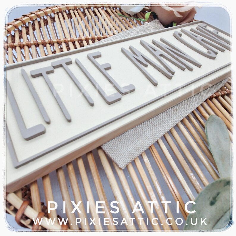 May include: A rectangular wooden sign with the words "LITTLE MAN CAVE" in raised, gray letters. The sign is set against a light background and framed in a light wood. The sign is placed on a woven surface.