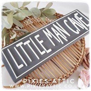 May include: A rectangular gray wooden sign with the words "LITTLE MAN CAVE" in white, raised lettering. The sign is set against a woven wicker background with decorative greenery and a pink flower. The sign is a decorative item.