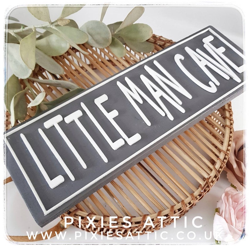 May include: A rectangular gray wooden sign with the words "LITTLE MAN CAVE" in white, raised lettering. The sign is set against a woven wicker background with decorative greenery and a pink flower. The sign is a decorative item.