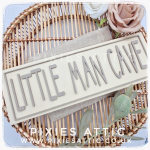 May include: A rectangular sign with the words "LITTLE MAN CAVE" in gray letters. The sign is on a light beige background, resting on a burlap cloth, and set on a woven wicker tray. The sign also has the text "PIXIES ATTIC" and "WWW.PIXIESATTIC.CO.UK".