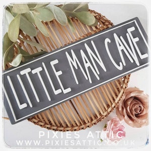 May include: A rectangular gray sign with the words "LITTLE MAN CAVE" in white lettering. The sign is placed on a woven basket with decorative flowers and greenery. The sign is a decorative item.
