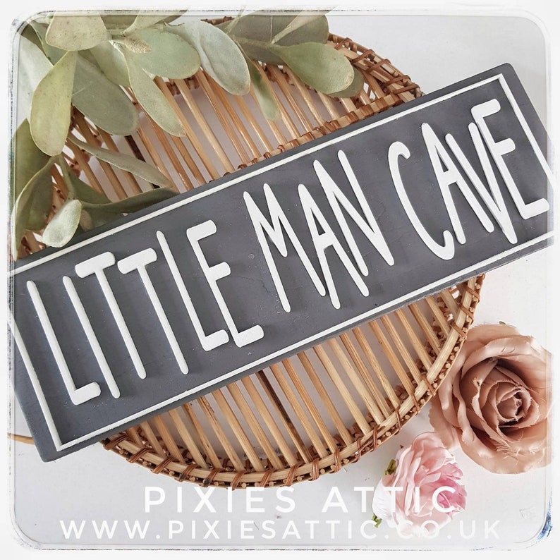 May include: A rectangular gray sign with the words "LITTLE MAN CAVE" in white lettering. The sign is placed on a woven basket with decorative flowers and greenery. The sign is a decorative item.