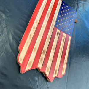 May include: Wooden American flag with red and white stripes and a blue field with stars. The flag has a rustic, distressed appearance with burnt edges and a unique shape. A patriotic home decor item.