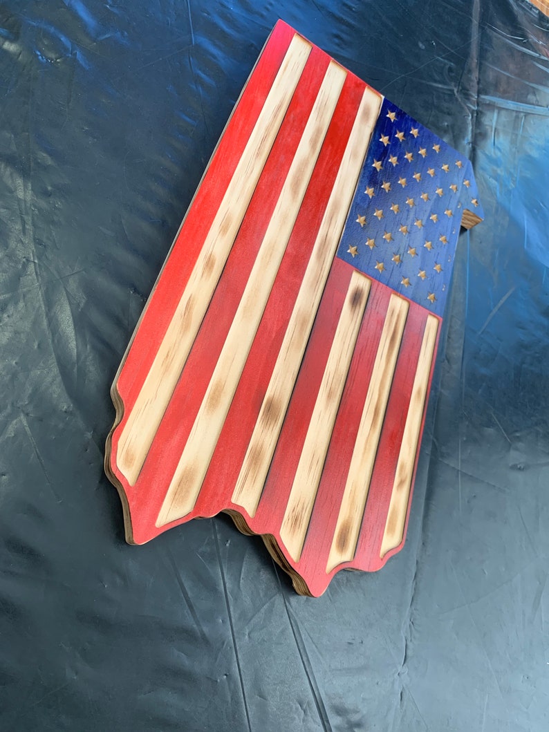 May include: Wooden American flag with red and white stripes and a blue field with stars. The flag has a rustic, distressed appearance with burnt edges and a unique shape. A patriotic home decor item.