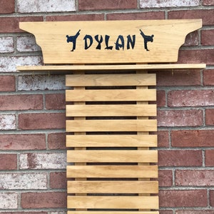 May include: Wooden karate belt display with the name "DYLAN" in dark blue lettering. The display also features two karate figures. The display is mounted on a red brick wall.
