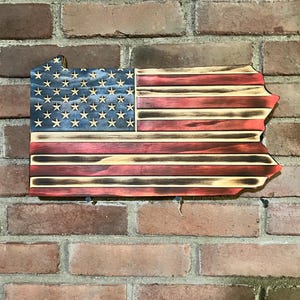 May include: Wooden wall hanging in the shape of Pennsylvania, featuring the American flag design. The flag has a blue field with white stars and red and white stripes. The wood has a distressed, burnt finish.