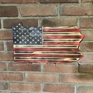 May include: Wooden wall art shaped like the state of Pennsylvania, featuring the American flag design. The flag has a blue field with white stars and red and white stripes. The wood has a distressed, burnt finish.
