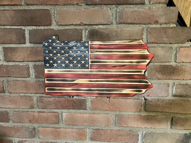 May include: Wooden wall art shaped like the state of Pennsylvania, featuring the American flag design. The flag has a blue field with white stars and red and white stripes. The wood has a distressed, burnt finish.
