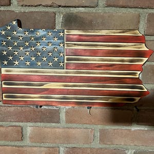 May include: Wooden wall art shaped like the state of Pennsylvania, featuring the American flag design. The flag has a blue field with white stars and red and white stripes. The wood has a distressed, burnt finish.
