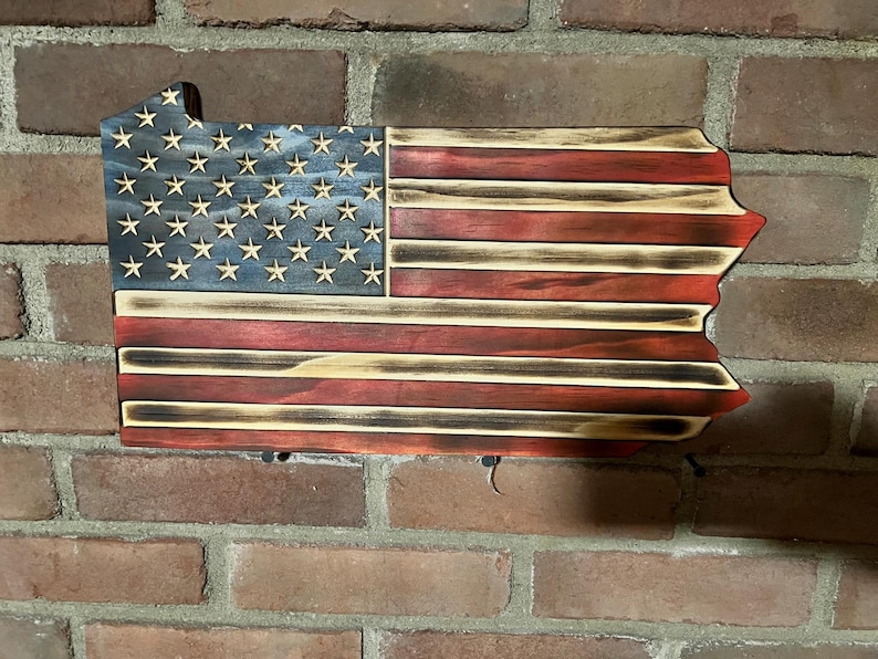 May include: Wooden wall art shaped like the state of Pennsylvania, featuring the American flag design. The flag has a blue field with white stars and red and white stripes. The wood has a distressed, burnt finish.