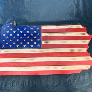 May include: Wooden wall art shaped like the state of Pennsylvania, featuring the American flag design. The blue section has white stars, and the stripes are red and natural wood. The wood has a slightly burned edge.