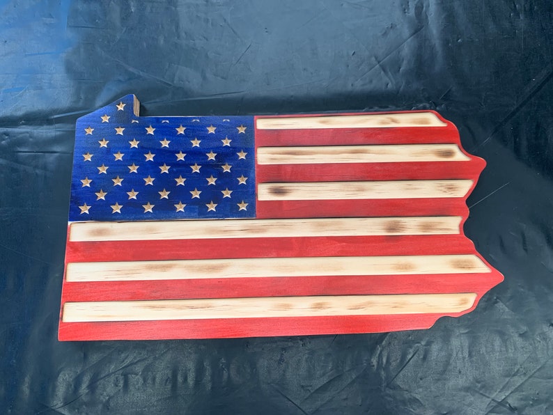 May include: Wooden wall art shaped like the state of Pennsylvania, featuring the American flag design. The blue section has white stars, and the stripes are red and natural wood. The wood has a slightly burned edge.