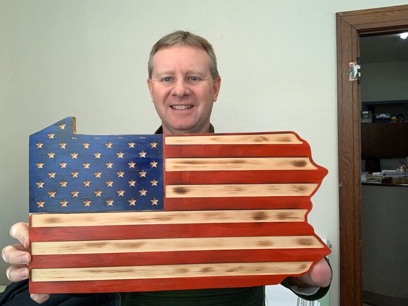 May include: A wooden sign in the shape of Pennsylvania, featuring the American flag design. The blue section has gold stars, and the red and white stripes are visible. The sign is made of wood and has a rustic, distressed look.