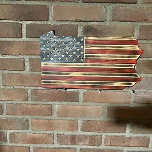May include: Wooden wall hanging in the shape of Pennsylvania, featuring the American flag design. The flag has a blue field with white stars and red and white stripes. The wood has a distressed, burnt finish.