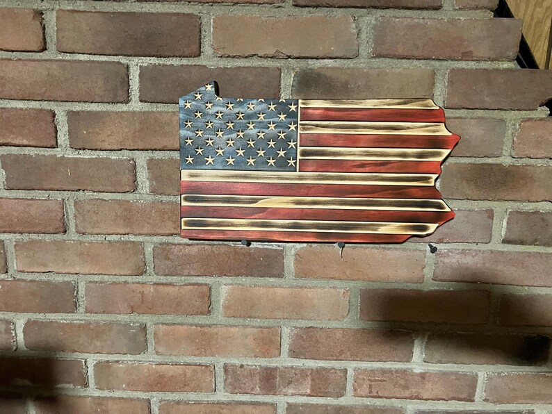 May include: Wooden wall hanging in the shape of Pennsylvania, featuring the American flag design. The flag has a blue field with white stars and red and white stripes. The wood has a distressed, burnt finish.