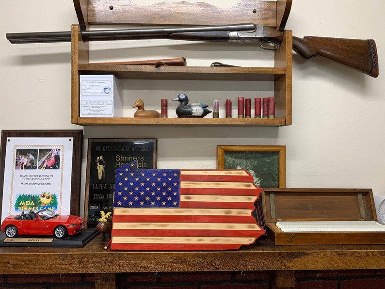 May include: A wooden shelf displays a double-barreled shotgun, a wooden duck decoy, and several shotgun shells. Below, a wooden Pennsylvania state flag, a red toy car, and framed plaques are visible.