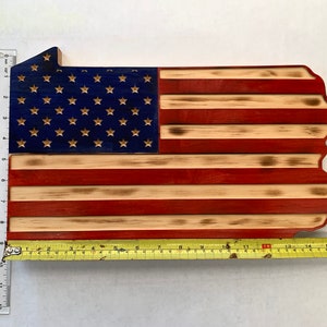May include: Wooden wall art in the shape of Pennsylvania, featuring the American flag design. The blue section has gold stars, and the stripes are red and natural wood. The piece measures approximately 30 inches long.