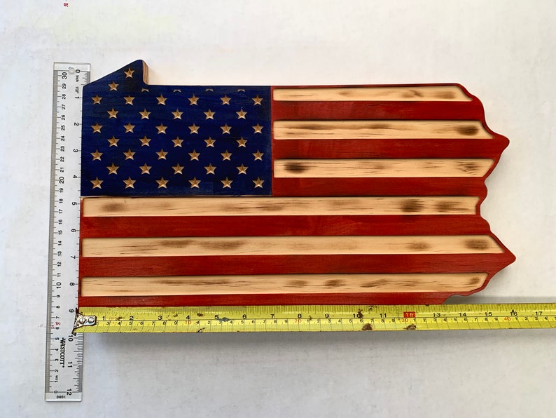 May include: Wooden wall art in the shape of Pennsylvania, featuring the American flag design. The blue section has gold stars, and the stripes are red and natural wood. The piece measures approximately 30 inches long.