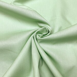 Könnte beinhalten: Nahaufnahme eines hellgrünen Stoffes mit glatter Textur. Der Stoff ist gefaltet und drapiert und erzeugt weiche Falten und Kurven.