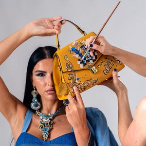 Könnte beinhalten: Senfgelbe Handtasche mit oberem Griff und Klappenverschluss. Die Tasche hat ein gemaltes Design mit den Worten "MAKE QUEEN" und einem dekorativen Schloss. Die Tasche wird von zwei Personen gehalten.