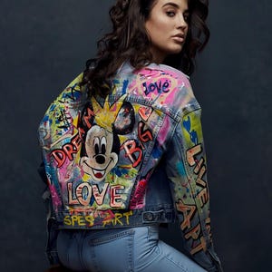 Könnte beinhalten: Hellblaue Jeansjacke mit farbenfrohen Graffiti-Kunstwerken. Auf der Rückseite befindet sich eine Mickey Mouse-Grafik und die Worte "DREAM BIG" und "LOVE". Die Ärmel sind mit "LIVE ART" und anderen künstlerischen Elementen verziert. Die Jacke wird mit hellblauen Jeans kombiniert.