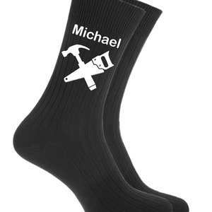 Pode incluir: Um par de meias pretas caneladas. As meias apresentam um gráfico branco de um martelo e uma serra cruzados, com o nome "Michael" acima das ferramentas. As meias são feitas de um material macio.