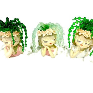 May include: Three decorative fairy planters with closed eyes, each featuring a floral crown and cascading green beaded hair. The fairies wear pink or white tops. These planters are ideal for small succulents or air plants.