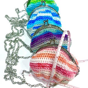 May include: Four colorful crochet coin purses with silver chain straps are displayed. The purses are in shades of green, blue, purple, and pink. A measuring tape is draped over the pink purse, showing its size.
