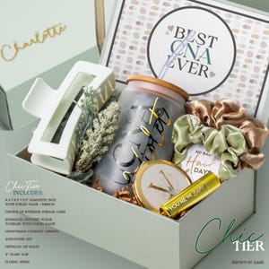 May include: A sage green gift box contains a frosted tumbler, compact mirror, scrunchie, lip balm, claw clip, and floral sprig. The tumbler has a gold foil name and straw. The box includes a magnetic closure and a ribbon. The box is labeled "Chic Tier".