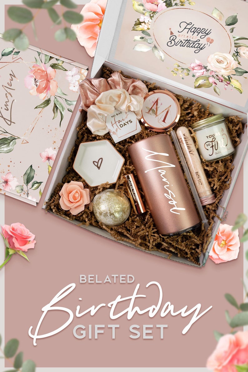 May include: A pink and white gift box filled with a variety of items, including a tumbler, bath bomb, and a candle. The box is decorated with a floral pattern and the words "Happy Belated Birthday".