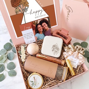 May include: A pink gift box containing a personalized tumbler with the name Lucy, a white jewelry case, a bath bomb, a tube of bath salts, a gold-tone compact mirror, a hair claw clip, a champagne-colored scrunchie, and a birthday card with a photo.