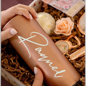 May include: A rose gold tumbler with the name "Raquel" in white script is held in a gift box filled with decorative paper. The box contains a bath bomb, a rose-shaped soap, and other gifts. Text on the image reads "Get the Full Picture" and "Boxed Gift Sets Available".