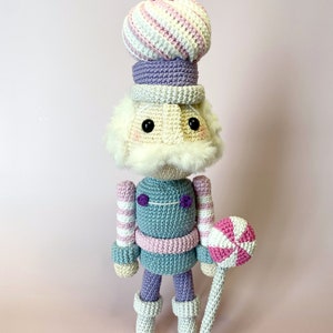 May include: A crocheted nutcracker doll with a pink and white striped hat, a purple hat, and a pink and white lollipop. The doll is wearing a blue and pink striped outfit and white boots.