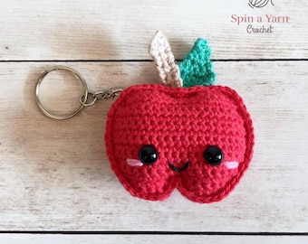 May include: A red crocheted apple keychain with a green and beige leaf on top. The apple has a smiling face with black eyes and pink cheeks. The keychain is attached to a silver key ring.