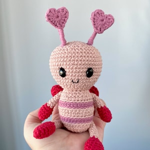 May include: A pink crocheted amigurumi toy shaped like a butterfly with large pink heart-shaped antennas and red wings. The toy has black eyes and a smiling mouth.