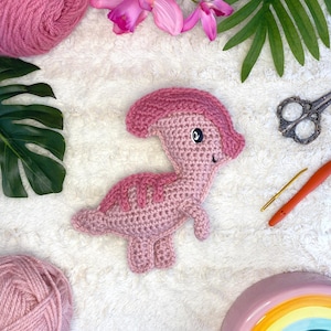 May include: A pink crocheted dinosaur toy with black eyes and a smiling mouth. The toy is sitting on a white fluffy surface.